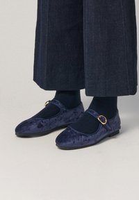 Bailarinas de terciopelo azul marino con diseño de Mary Jane, que presentan una puntera redonda, una correa al tobillo con un cinturón en tono dorado y una suela plana, llevadas con calcetines oscuros.