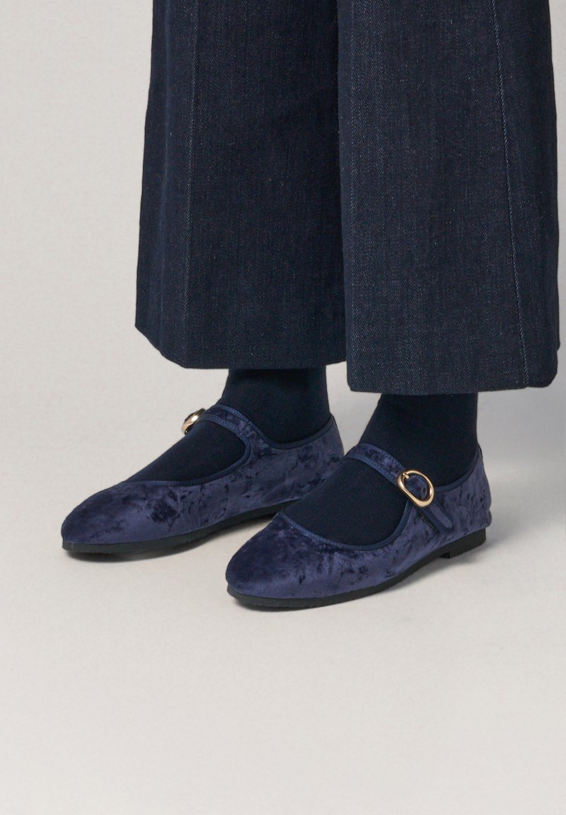 Bailarinas de terciopelo azul marino con diseño de Mary Jane, que presentan una puntera redonda, una correa al tobillo con un cinturón en tono dorado y una suela plana, llevadas con calcetines oscuros.