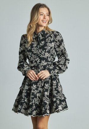 Vestido negro con bordado floral blanco, con cuello de solapa, mangas largas y un diseño de volantes en capas en el dobladillo.