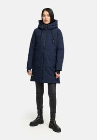 Kvinde står iført en navyblå hættetrøje vinterjakke, sorte skinny jeans og sorte chunky støvler mod en ensfarvet lys baggrund.