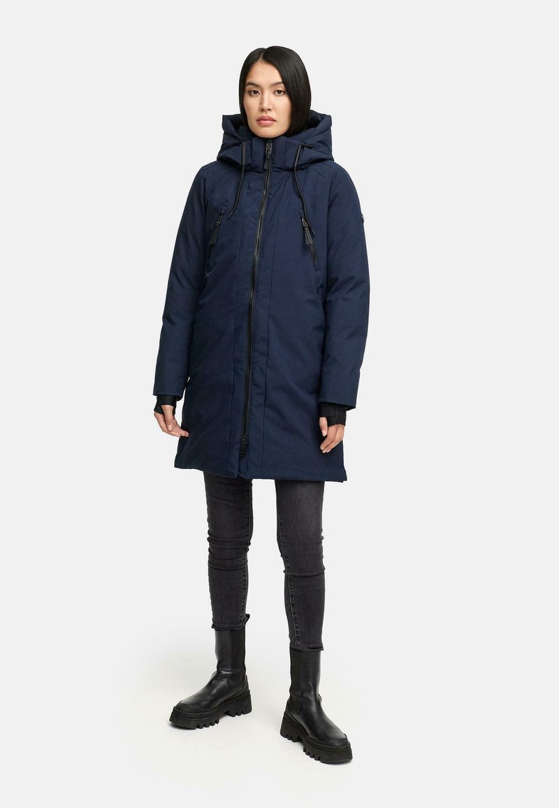 Donna in piedi che indossa un cappotto invernale con cappuccio blu navy, jeans skinny neri e stivali neri chunky su uno sfondo chiaro e semplice.