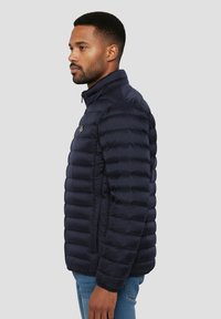 Veste matelassée bleu marine avec coutures horizontales, col montant et fermeture éclair à l'avant. Comprend des poches latérales et un petit emblème sur la poitrine.