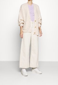 Beige gestrickte Strickjacke mit Knöpfen, kombiniert mit einem helllila Rüschenhemd und cremefarbenen, weit geschnittenen Hosen. Weiße Sneakers vervollständigen das Outfit.