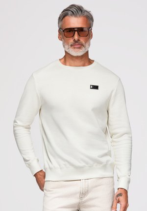 Homme aux cheveux gris et à la barbe portant des lunettes de soleil marron, un sweat-shirt blanc à col rond et un pantalon clair, debout avec une main dans la poche.