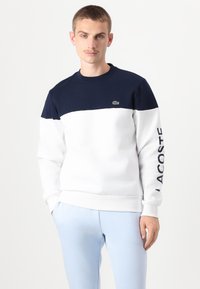 Krāsaini blokota sudraba krekls, izgatavots no kokvilnas, ar tumši zilu un balto krāsu daļām, apaļu kaklu un "LACOSTE" tekstu uz piedurknes.