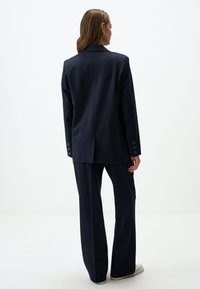 Blazer blu navy sagomato con spacchi singoli, abbinato a pantaloni a gamba larga. Entrambi realizzati in tessuto morbido con dettagli in bottoni sulle maniche.