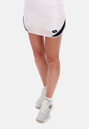 Femme portant une jupe de sport blanche avec des accents noirs et des chaussures de sport blanches, debout les jambes croisées devant un fond uni.