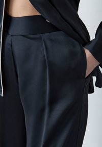 Zwarte satijnen broek met een gladde textuur, voorzien van een brede tailleband en een geplooide voorkant. Een hand rust in de zijzak, wat een casual uitstraling toont.