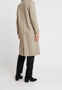 Long plaid coat in beige with brown accents, featuring a straight cut, long sleeves, and back vent, paired with black pants and block-heeled shoes.