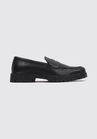 Mocasines de cuero negro con punta redonda, suela de goma gruesa y una tira decorativa en la parte frontal. Textura suave, diseño minimalista.
