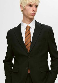 Czarny garnitur o dopasowanej sylwetce, z pojedynczym zapięciem z przodu i dwoma guzikami; zestawiony z białą koszulą i pomarańczowym krawatem w paski.