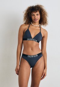 Temno siva bikinija s kompletom s trikotno obliko in visokim pasom. Vključuje logotip Calvin Klein na elastičnih trakovih. Gladka tekstura tkanine.