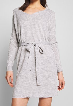 Frau trägt ein hellgraues, langärmeliges Strickkleid mit V-Ausschnitt und gebundenem Gürtel in der Taille.