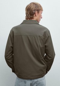 Khaki jakke med krave, glat tekstur og lange ærmer. Ryggen har en vandret søm på tværs af skuldrene.