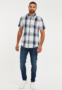 Camisa de cuadros de manga corta en blanco, azul marino y gris. Presenta cierre de botones, cuello y pequeño logo en el pecho. Combinada con jeans ajustados.