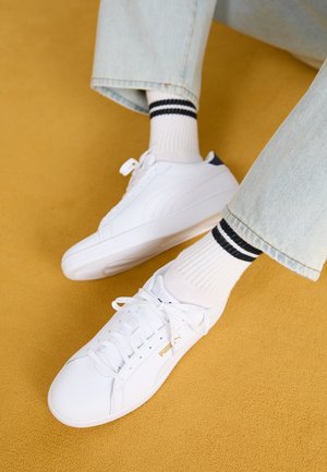 Baskets blanches avec une tige en cuir lisse, un design à lacets et un accent logo doré. Associées à des chaussettes blanches côtelées avec des rayures noires.