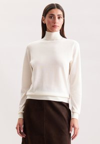 Kremowy sweter w formie golfu z lekkiego materiału, z długimi rękawami i prążkowanymi mankietami przy dolnej krawędzi. Zestawiony z brązową spódnicą z zamszu.