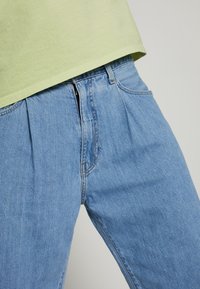 Jeans denim berwarna biru muda dengan lipatan depan, dilengkapi ritsleting dan kaki yang sedikit menyempit. Dipadukan dengan kemeja lengan pendek berwarna hijau muda.