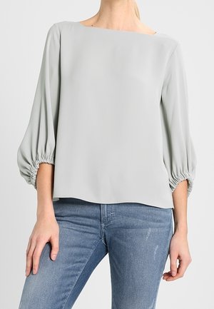 Blusa grigia chiara con scollatura quadrata e maniche lunghe a sbuffo raccolte ai polsi. Tessuto liscio con vestibilità rilassata, abbinata a denim.