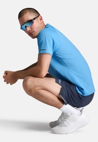 Mees kükitab, seljas helesinine lühikeste varrukatega särk, tumedad lühikesed püksid ja valged tossud. Tal on peegeldavad päikeseprillid ja lühike soeng.