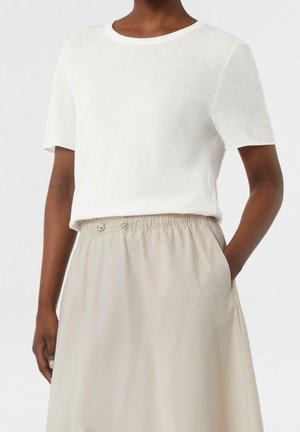 Kvinna iförd en enfärgad vit T-shirt med kort ärm, nedstoppar i en högmidjad beige kjol med elastiskt midjeband och fickor.