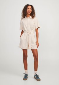 Vestido beige de manga corta con botones y cinturón anudado, dos bolsillos laterales y un bolsillo en el pecho, hecho de una tela texturizada.