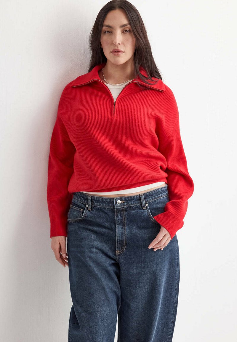 Femme debout contre un mur blanc, portant un pull rouge zippé et un jean bleu foncé ample.