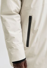 Beebitooni lähedalt vaadatuna beežist jope varrukas, millel on must vertikaalne tõmblukuga tasku ja tumedanahaline käsi, mis on osaliselt nähtav varruka ääres.