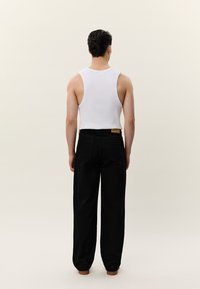 Άνδρας που στέκεται με την πλάτη στραμμένη, φοράει λευκό αμάνικο μπλουζάκι και φαρδιά μαύρη παντελόνα με ψηλή μέση, σε απλό ανοιχτό φόντο.