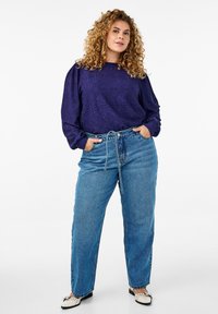 Purpura acu caurumu tops ar puffētām piedurknēm, kombinēts ar augsto jostasvietu zilām džinsa biksēm. Apģērbs piedāvā sasietu vidukļa detaļu un plakanas kurpes.