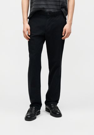 Osoba nosząca czarne spodnie o prostych nogawkach, czarne skórzane mokasyny oraz włożoną do środka pasiastą koszulę, stojąca na tle jednolitego tła.