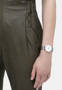 Silberne Metall-Uhr mit einem weißen Zifferblatt, getragen an einer Hand, neben einem olivgrünen Ledermantel mit sichtbaren Nahtdetails und Textur.