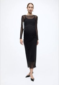 Fekete ruha hosszú, áttetsző ujjakkal, ráncolt részletekkel, térdig érő, apró csillogó díszítéssel, fekete hegyes orrú cipővel párosítva.