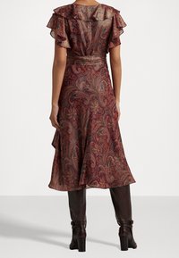 Schwingendes, gemustertes Kleid in sattem Burgunderrot mit Paisley-Design. Verfügt über Rüscheärmel und eine taillierte Taille, kombiniert mit braunen knielangen Stiefeln.