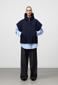 Granatowa bezrękawnik z zamkiem i kapturem, noszony na jasnoniebieskiej koszuli. Model ma na sobie ciemne spodnie w szerokie nogawki i trzyma ręce w kieszeniach.