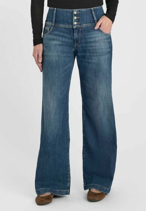 Femme portant un jean bleu taille haute à jambes larges avec trois boutons visibles et des chaussures marron, debout avec une main dans la poche.