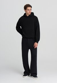 Felpa nera con tasca frontale, vestibilità comoda. Abbinata a pantaloni neri a gamba larga. Entrambi realizzati in tessuto morbido, con vita regolabile tramite cordino.