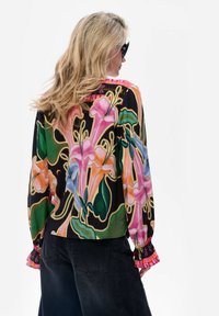 Kobieta z blond włosami nosząca czarne okulary przeciwsłoneczne i kolorową, kwiatową bluzkę w odcieniach różu, pomarańczu, niebieskiego i zielonego, w połączeniu z czarnymi spodniami.