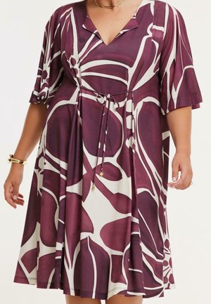 Mujer que lleva un vestido hasta la rodilla con estampado floral en púrpura y blanco, mangas cortas y cinturón anudado en la cintura.