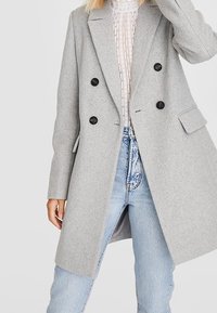 Cappotto doppiopetto grigio in misto lana con rever a punta, bottoni neri e tasche anteriori, indossato sopra una maglietta leggera e testurizzata e jeans blu.