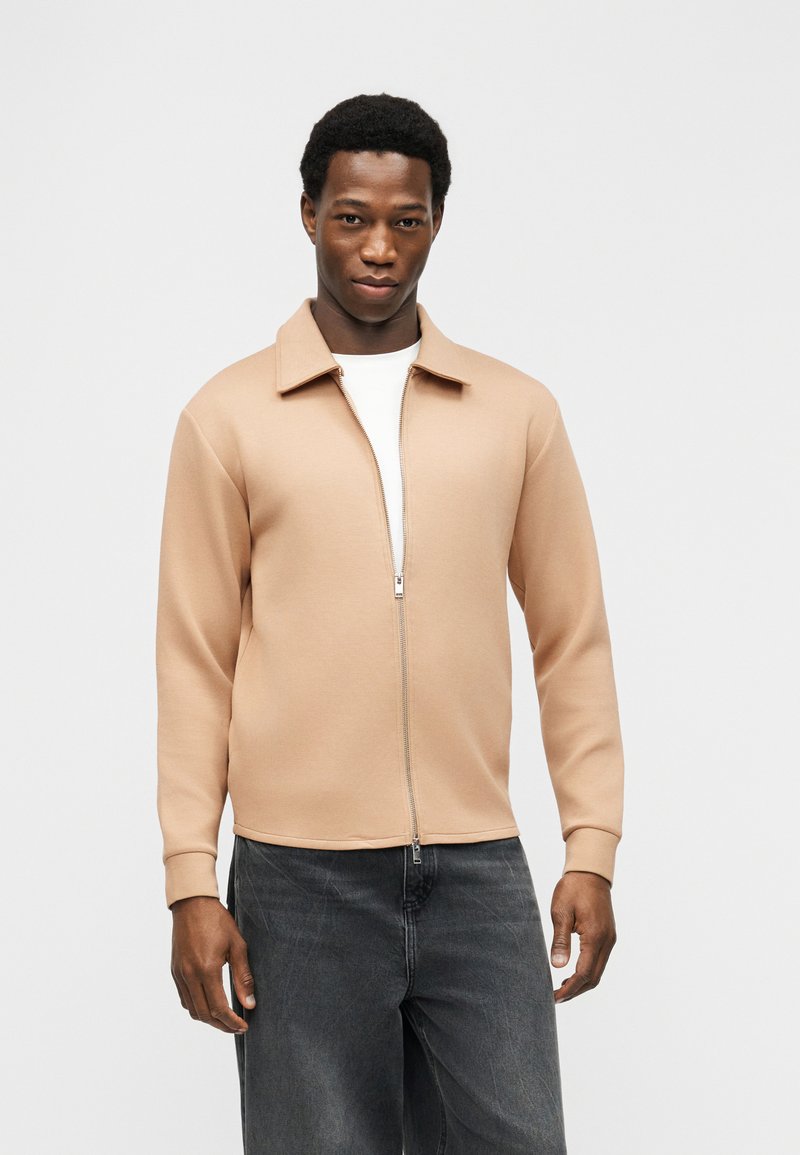 Άνδρας με μπεζ ζακέτα φερμουάρ πάνω από λευκό πουκάμισο και σκουρόχρωμο γκρι τζιν, στέκεται μπροστά σε απλό ανοιχτό γκρι φόντο.