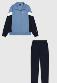 Giacca a track blu e blu navy con strisce bianche; chiusura con zip, polsini a costine. Pantaloni sportivi blu navy abbinati con vita elastica e logo.
