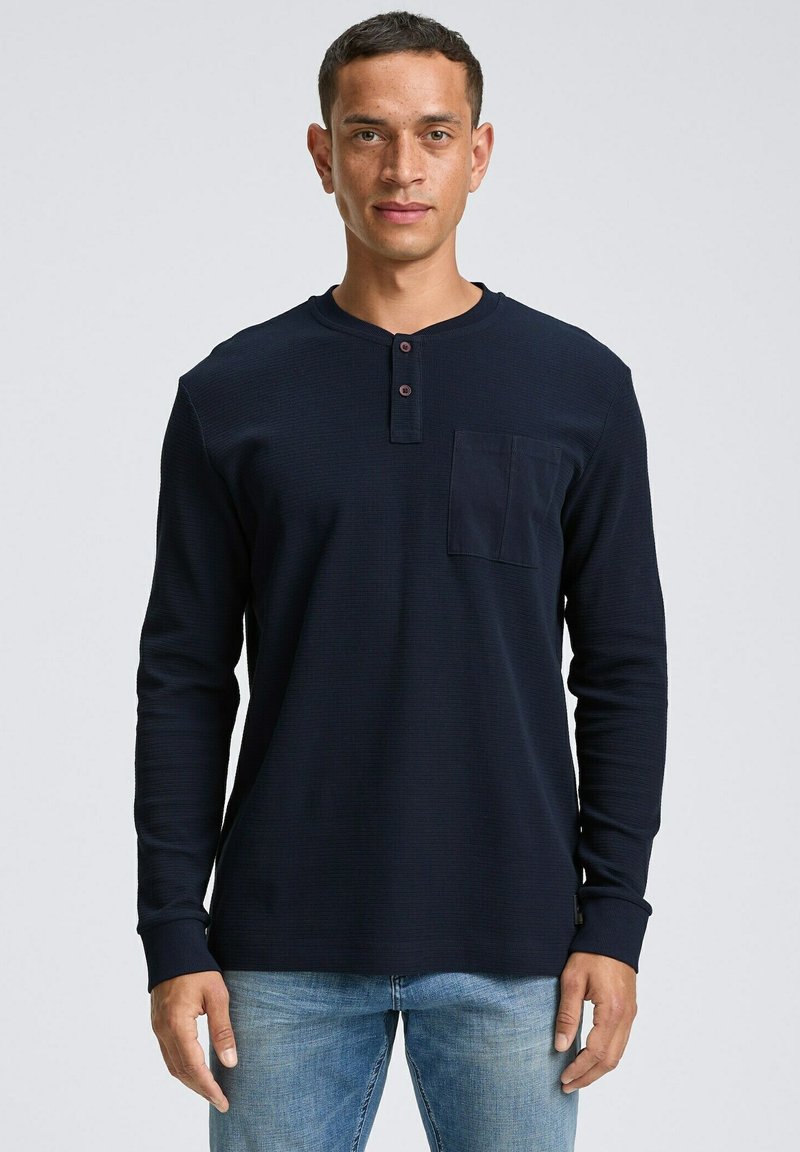 Marineblauwe longsleeve henleyshirt van textiel met drie knopen, voorzien van een borstzak en geribbelde manchetten. Gepaard met lichte jeans.