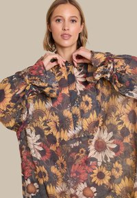 Bluse med lange ermer i gjennomsiktig stoff med blomstermønster som har solblomster, samt innslag av rødt og gult. Løs passform med rynkede ermer.