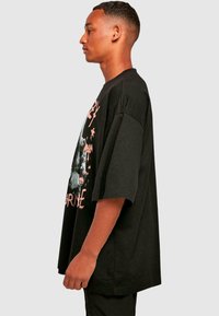Tricou oversized negru cu mâneci largi, prezentând un design grafic în roz și alb pe partea din față. Fabricat dintr-un material moale, cu o croială lejeră.