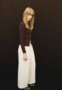 Cardigan marrón de manga larga con botones, combinado con pantalones anchos blancos y sandalias con tiras, sobre un fondo oscuro.