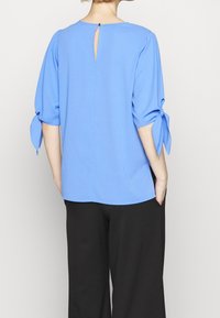 Blusa blu in tessuto morbido, con apertura a chiave sul retro, maniche a tre quarti con fiocchi e vestibilità morbida. Abbinata a pantaloni neri.