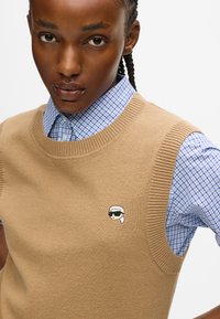 Zbliżenie osoby noszącej beżowy sweter z dzianiny z małym haftowanym logo w kształcie twarzy na niebieskiej koszuli w kratę z kołnierzykiem.