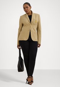 Blazer beige su misura con bottoni dorati e dettagli sulla tasca del petto, abbinato a jeans skinny neri e tacchi a strappo neri. Presenta una texture minimale.