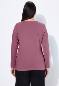 Langärmeliges, traubenfarbenes Baumwoll-T-Shirt mit Rundhalsausschnitt und glatter Textur, getragen mit schwarzen Hosen. Rückansicht zeigt ein einfaches Design.